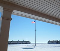 View of snow covered open area with flag pole in the middle and sandstone buildings in the background.