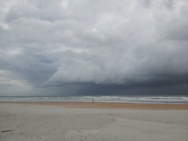 Menacing storm clouds turn the sky dark over a lone, small human figure on a wide beach
