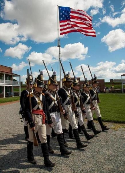 Fort McHenry Guard living history volunteers marching in U.S. Corps of Artillery uniforms. Star-Spangled Banner in background.