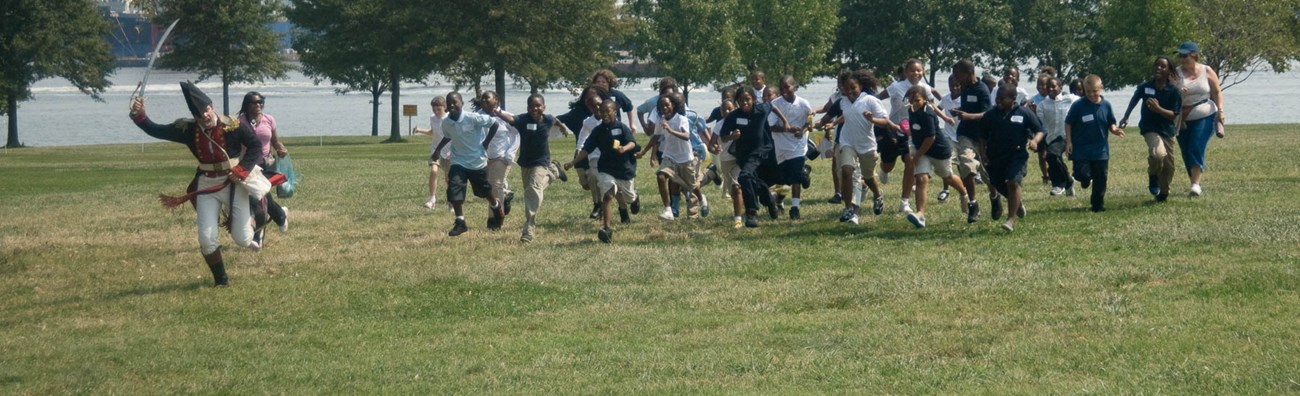 A living historian leads a group of kids running across a grass lawn.