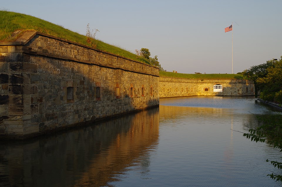 A view of the outside two stone bastion walls, a water moat, and a flagpole with the American flag flying against a blue sky.