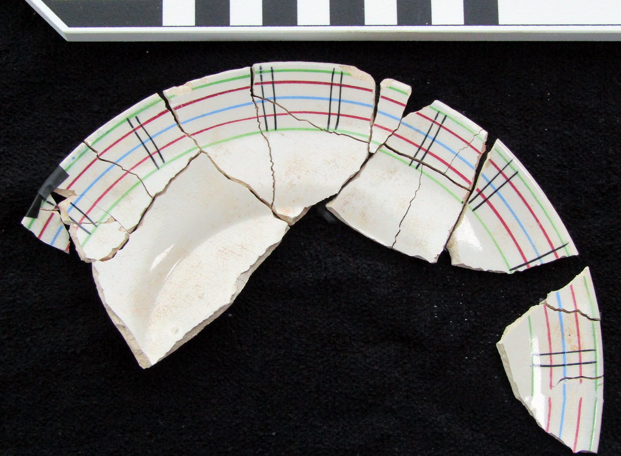 White plate with different colored thin lines around the edge. Multiple sherds have been put back together.