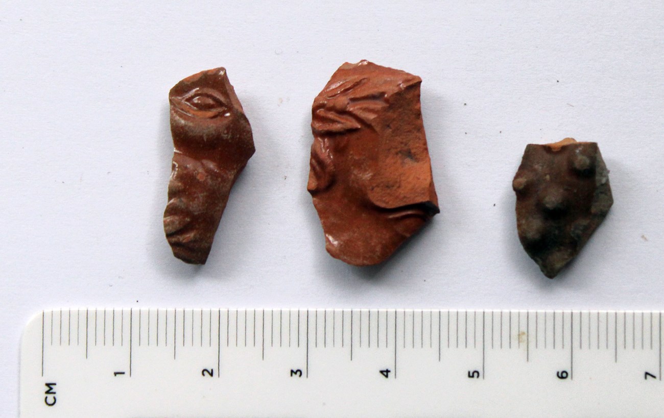 Three orange-red fragments of ceramic pipe bowl. Eyes are visible on two fragments, and the other has raised bumps.