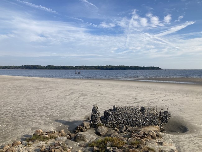 The stony remnants of Cockspur Island's historic north pier mark a sandy shoreline on a bright, cloudy day with the Savannah River in the background.