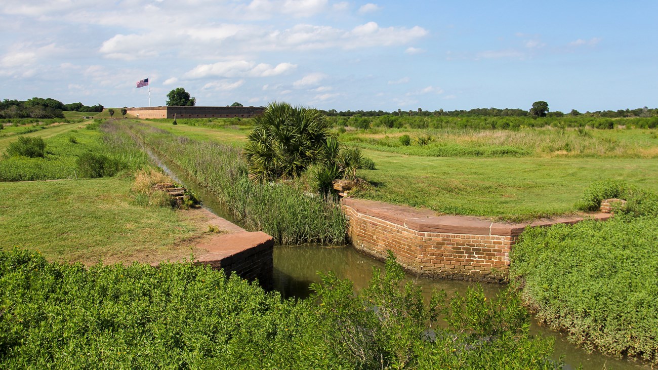 A sandstone a brick sluice gate ruin marks the beginning of the feeding canal. A red bricked Fort Pulaski flies a large U.S. in the background. A large expanse of marsh covers the way.