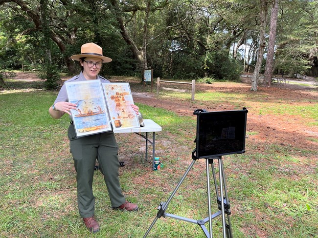 A ranger holds an image up while looking into an iPad. Behind her is a camera with more images and a metal helmet