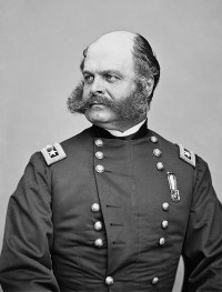 A man sitting in a military uniform with sideburns connecting to a large beard