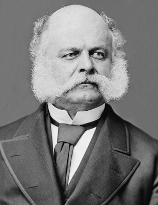 A photograph chest-up of a man with large all-white sideburns connected to his mustache. He's dressed in a suit and tie