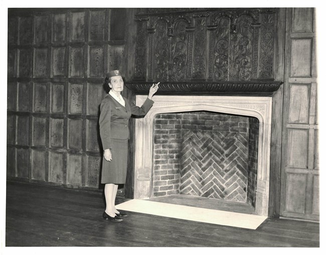 Black and white photo of female Park Ranger standing by Elizabethan Room fireplace.