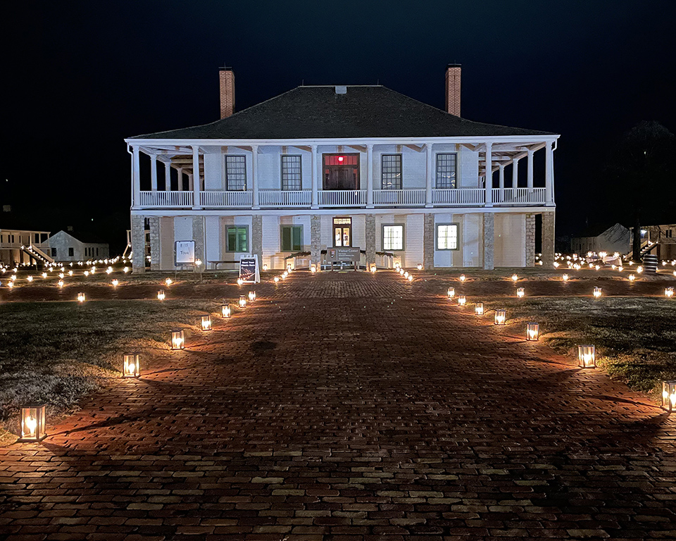 Image of a white building with stone pillars on the first floor and a white railing around the porch on the second. Candle lanterns line the sidewalk leading to the building.