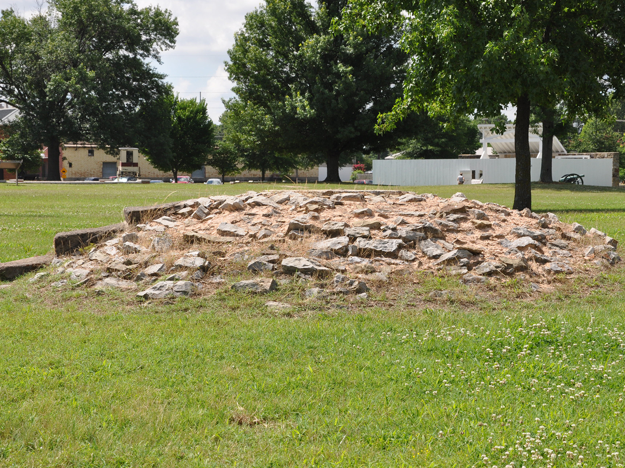 Three small cement stairs lead to the top of the mound of gray stones held together by tan mortar. The mound is surrounded by bright green grass on a sunny day.