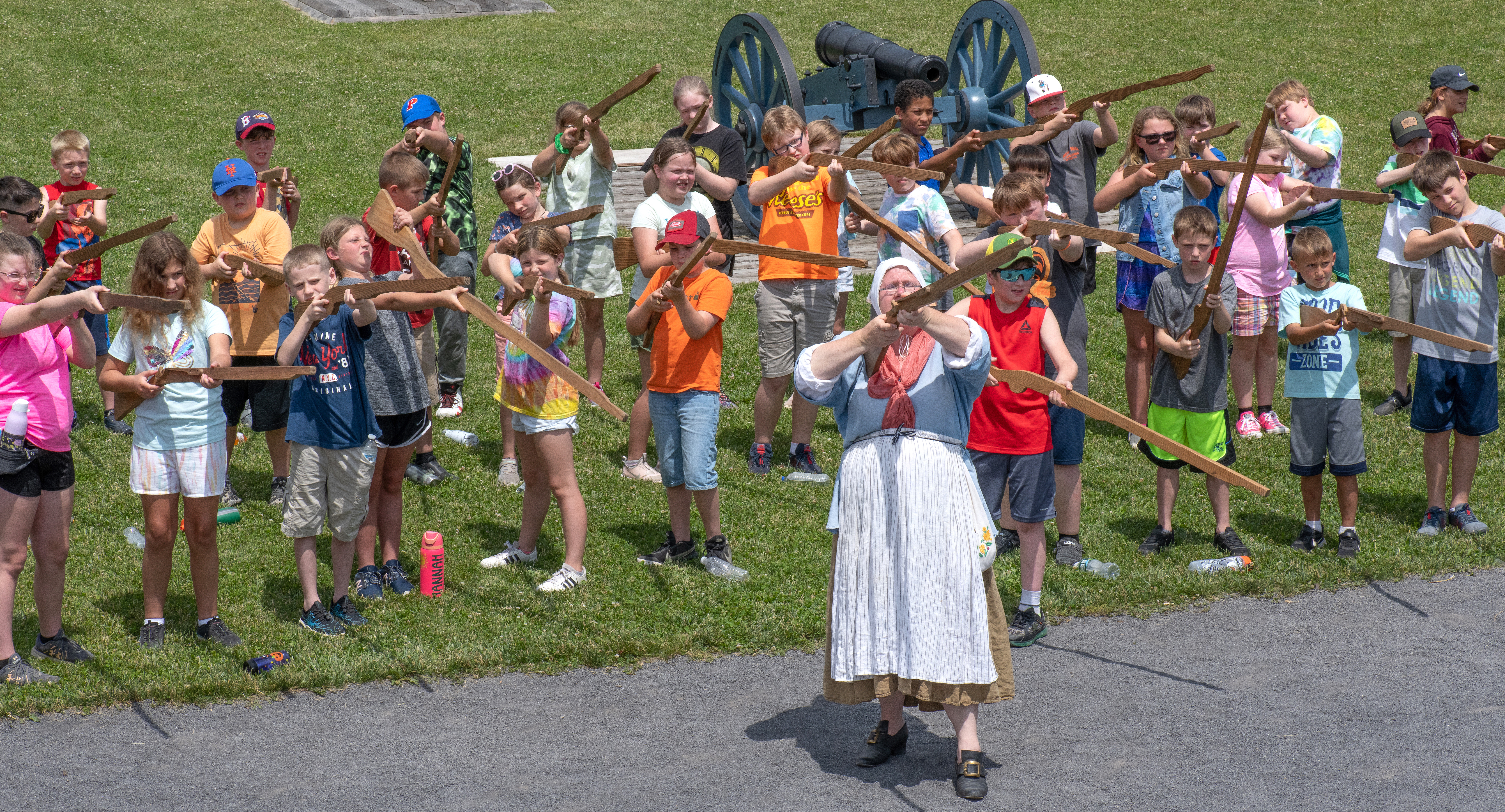 Dozens of children in lines behind a woman in 18th C clothing. They are all holding and pointing wooden muskets in the air.