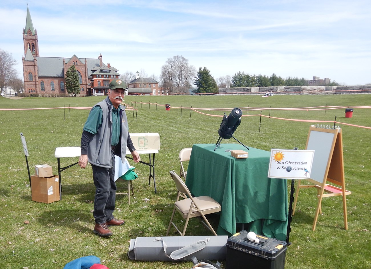 A man with a swanky mustache and hat moves between tables with various types of scientific equipment on them.