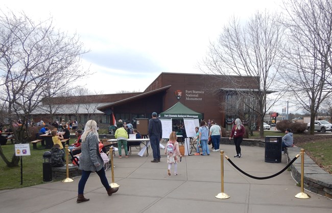 A crowd of people garther and move around a pavilion in front of a large wooden building with an NPS arrowhead on it.