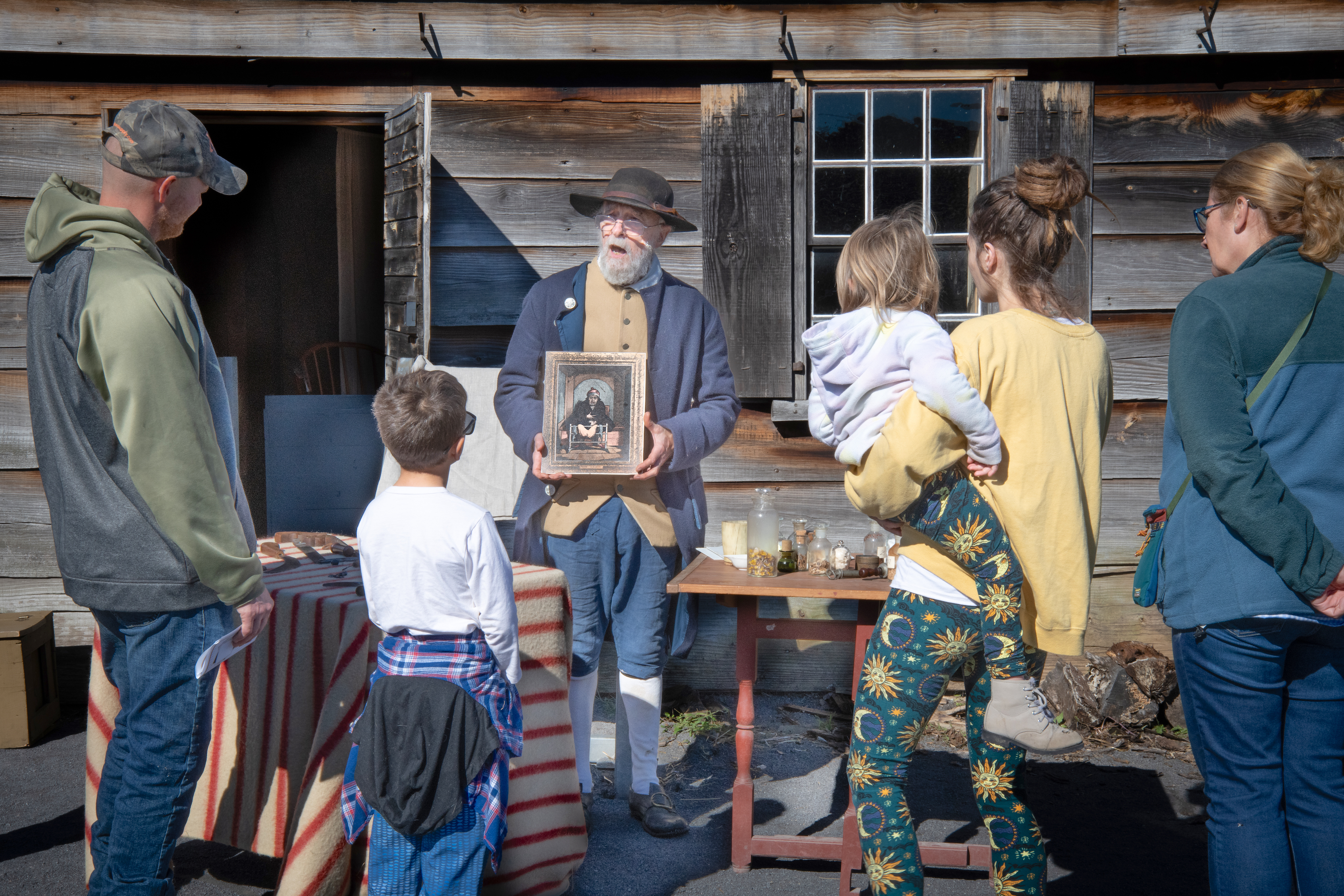 A man with beard in 18th C style clothing stands holding something in front of a family group.