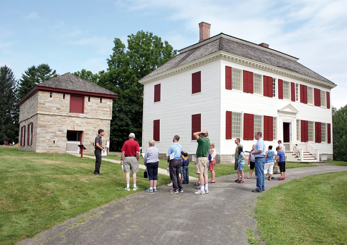 Johnson-Hall-State-Historic-Site-1200x850 a large two-story building with a shingled roof. It is painted white with a central doorway. two windows sit on each side of the doorway, and five windows spaced evenly across the second floor. The door and window shutters are dark red.