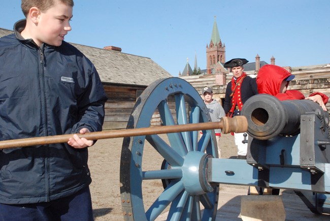 One child inserts a long stick into a muzzle of a cannon, while a second watches the breach, and a Continental soldier supervises.