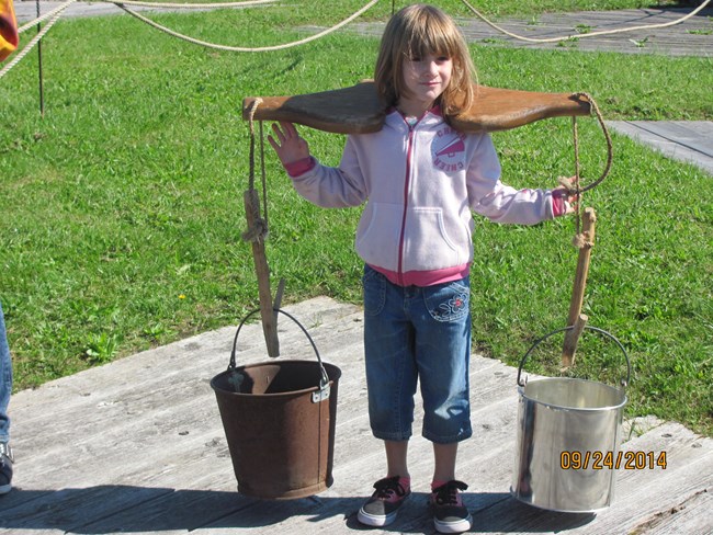 A little girl stands with a yolk and two buckets over her shoulder.
