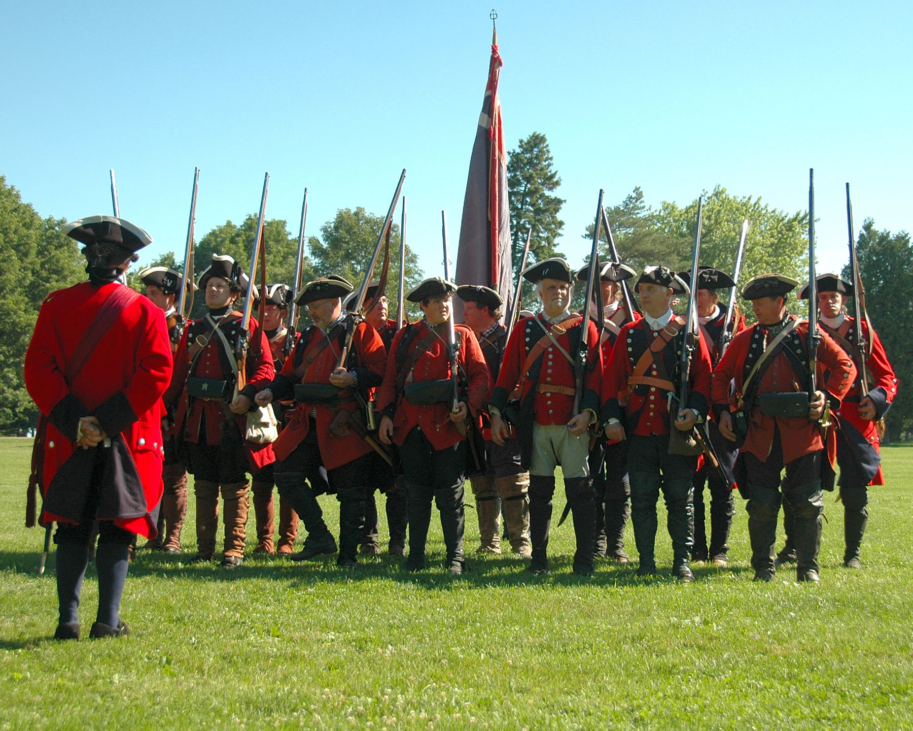 men in red jackets stand in a line with muskets.