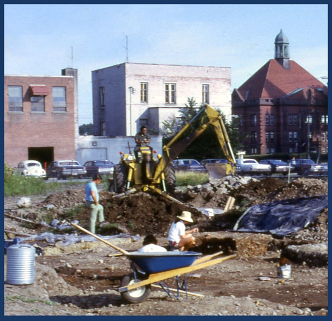 Wide view of the archeological excavations with tools as small as buckets and as large as a backhoe, buildings that were eventually demolished are in the background.