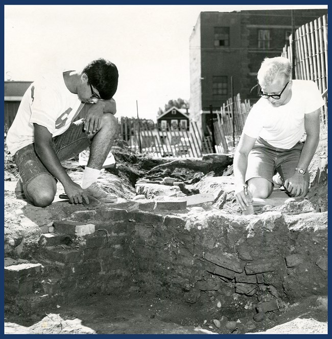 Two people sit and excavate with a trowel, uncovering a brick structure that was later determined to be the bakehouse.