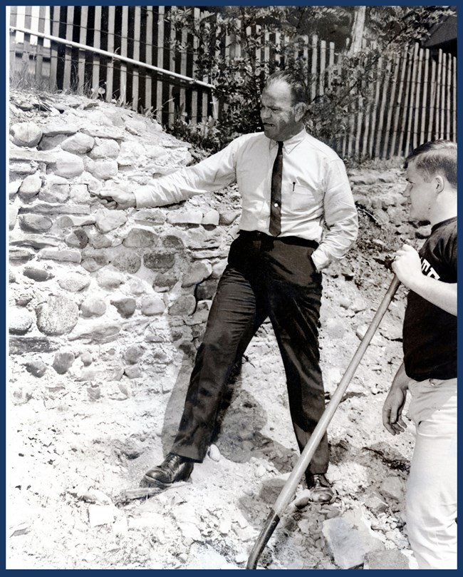 J. Duncan Campbell stands in a collared shirt and slacks smoking a cigar and pointing at an archeological profile.