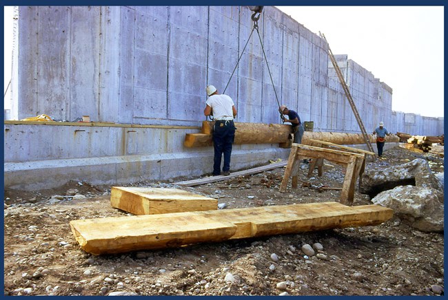 A large concrete wall towers over workers who are attaching a wood facade with the help of a crane.
