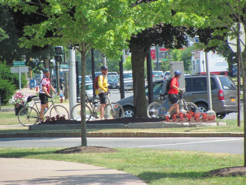3 cyclists ride in a line at an intersection. They wear neon colored clothing.