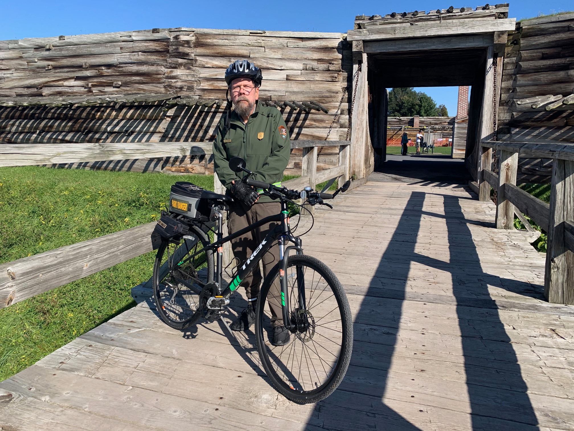 A park ranger stands next to a bicycle on a wooden bridge.