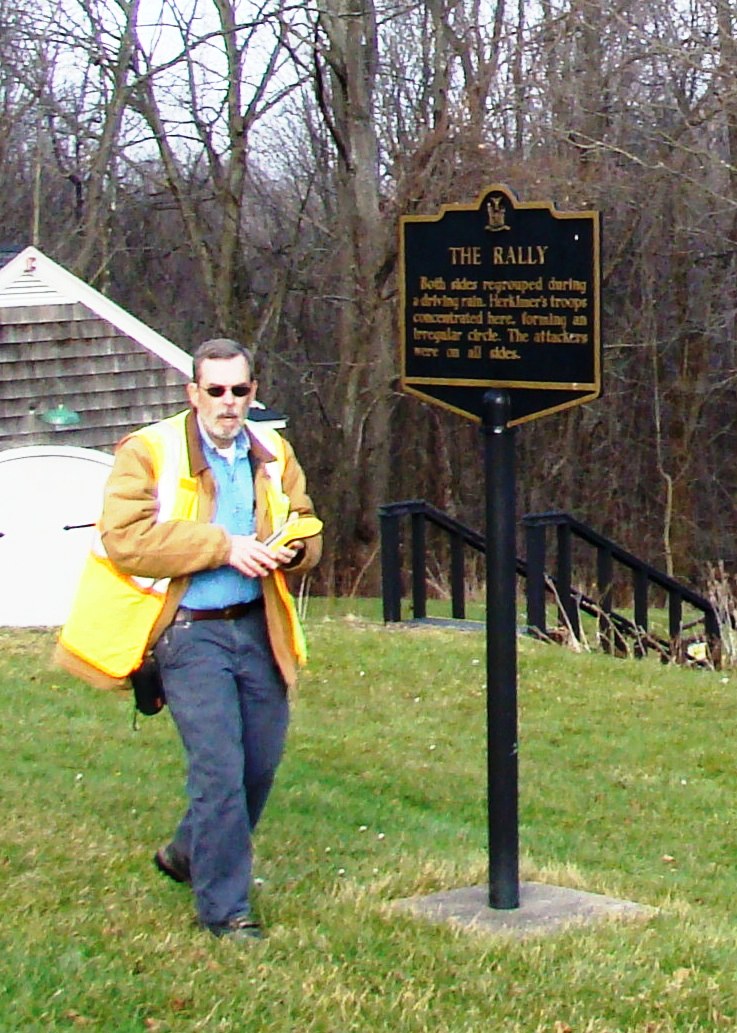 a man walks thorough a green field, past a gold and black sign