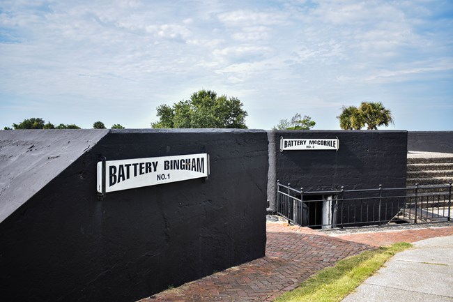 Two black batteries facing into Charleston Harbor. Behind them are palm trees on the beach. There is a red brick path in front of them