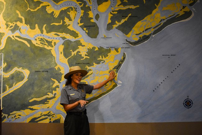 A female ranger dressed in a green and grey park uniform wearing a campaign hat stands in front of a large map of Charleston.
