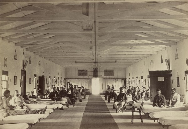 Black and White photo of a large, open, and sparsely decorated pavilion-style building with about 30 hospital beds, each occupied by a soldier, lining each side of the building.