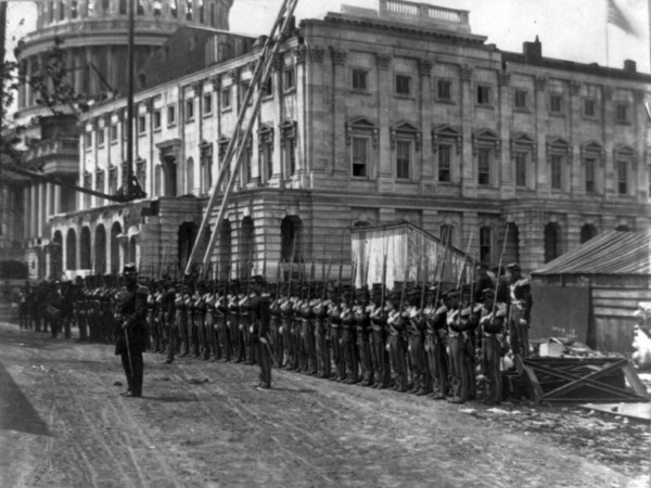 A large group of Union soldiers are seen standing at attention on a dirt road running immediately in front of the US Capitol. All the soldiers are seen equipped with rifles with the exception of their commanding officer standing in the foreground.