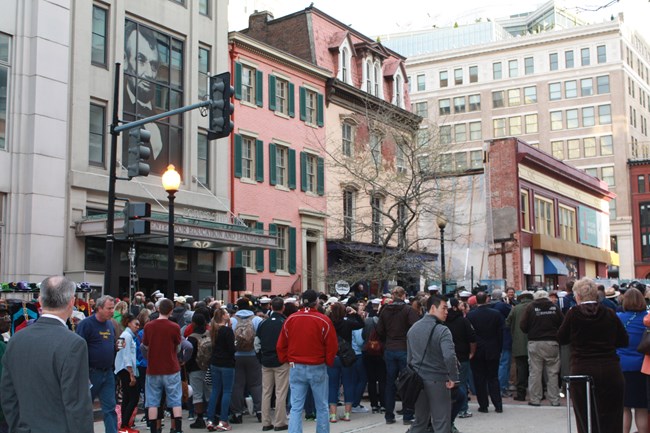 A crowd of people stands in the street outside a red brick house with green shutters.