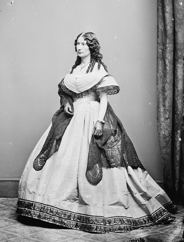 Black and white photograph of a white woman with dark curly hair standing in an elegant hooped dress with lace accessories
