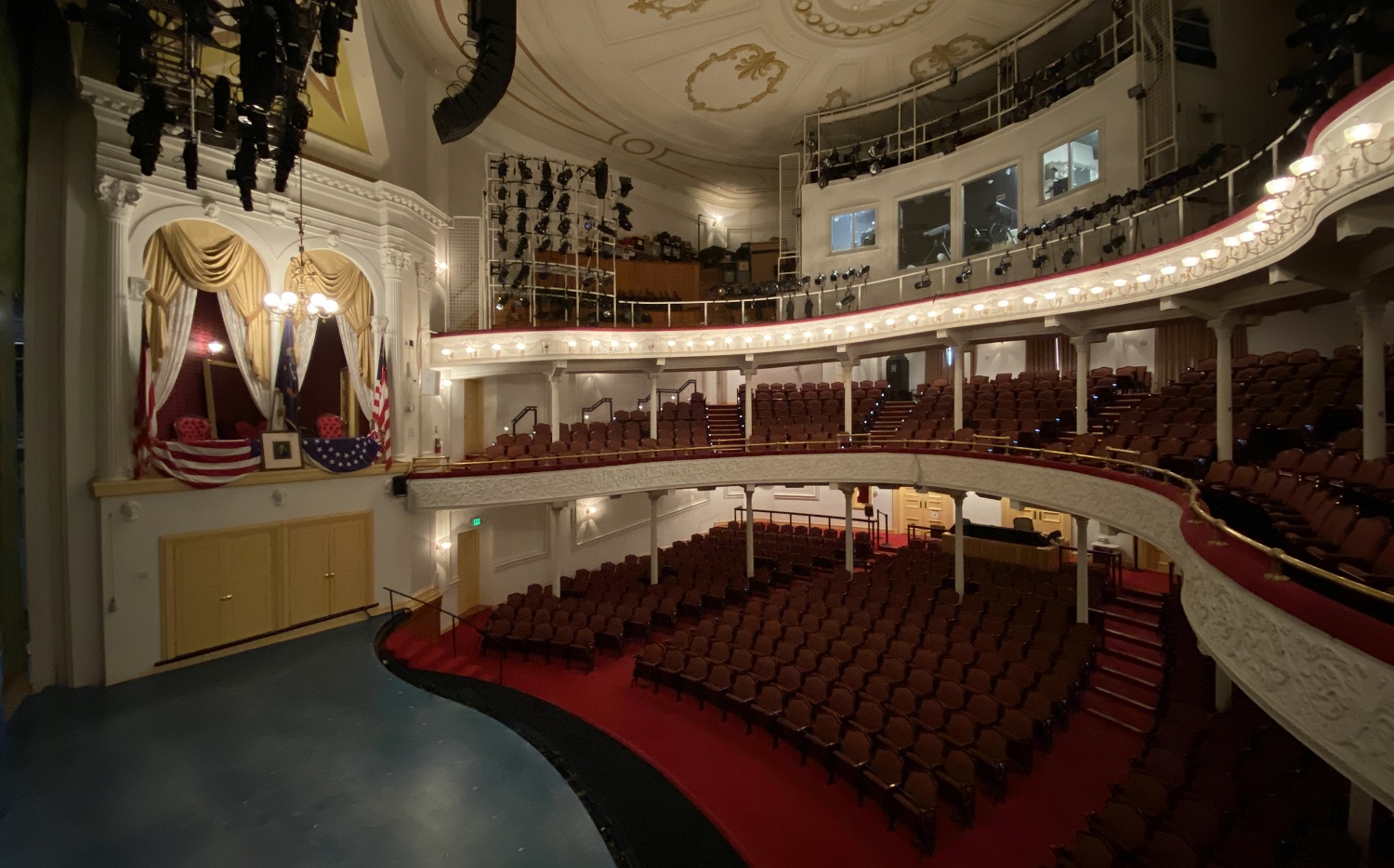 Photo of theatre stage and audience seating on three levels, with the box seats decorated with flags on the other side of the stage