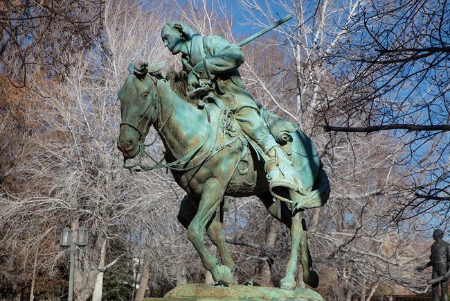 A bronze statue that has turned green of Kit Carson riding horse and carrying a rifle under is left arm