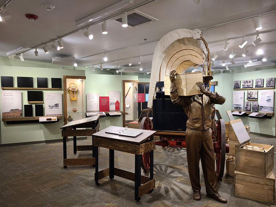 A mannquin of a man holding a crate on his shoulder standing in front of a covered wagon.