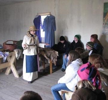 Woman in historic clothing consisting of a white skirt and to with a blue apron and straw hat talking to visitors in a historic structure with a wood floor and canvas walls.