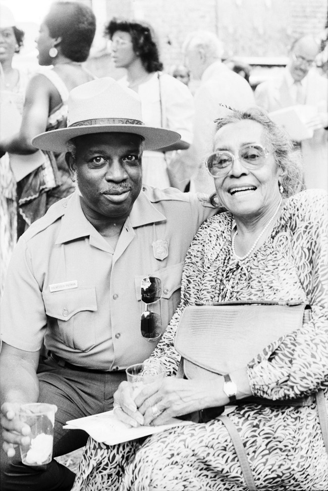 Black and white photo of an African American Ranger sitting with an African American woman in a dress both smiling with more people in the background.