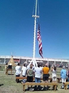American flag on white flagpole under a clear blue sky. Visitors stand under the flag.