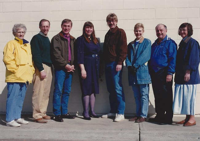 Eight people standing facing the camera smiling against a white brick wall.