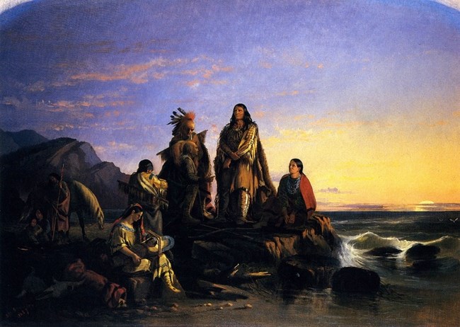 A group of American Indians some standing some sitting with a horse to the left of them and an ocean to the right.