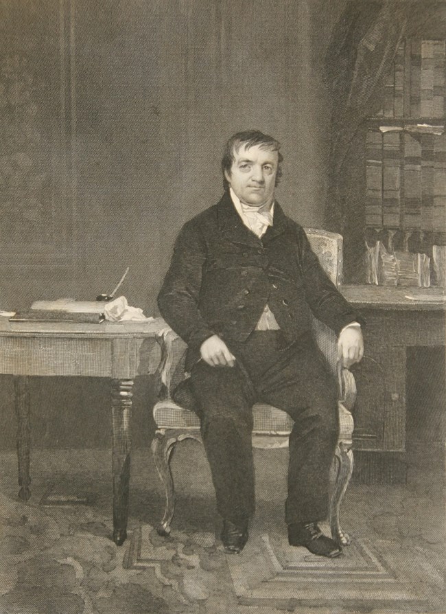 Black and white image of a man with short hair in a dark colored formal 1800s suit sitting next to a wooden desk in a library.
