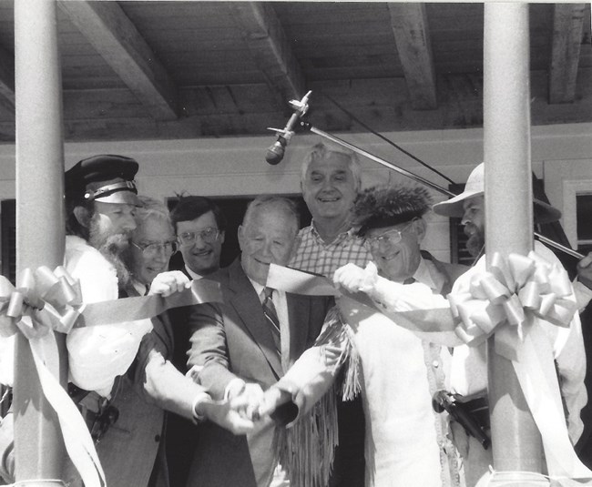 Seven men stand on porch cutting ribbon.