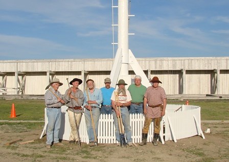 Seven men in historic dress some holding tools stand in front of white fence and large white flagpole with white walls behind and a blue sky above.