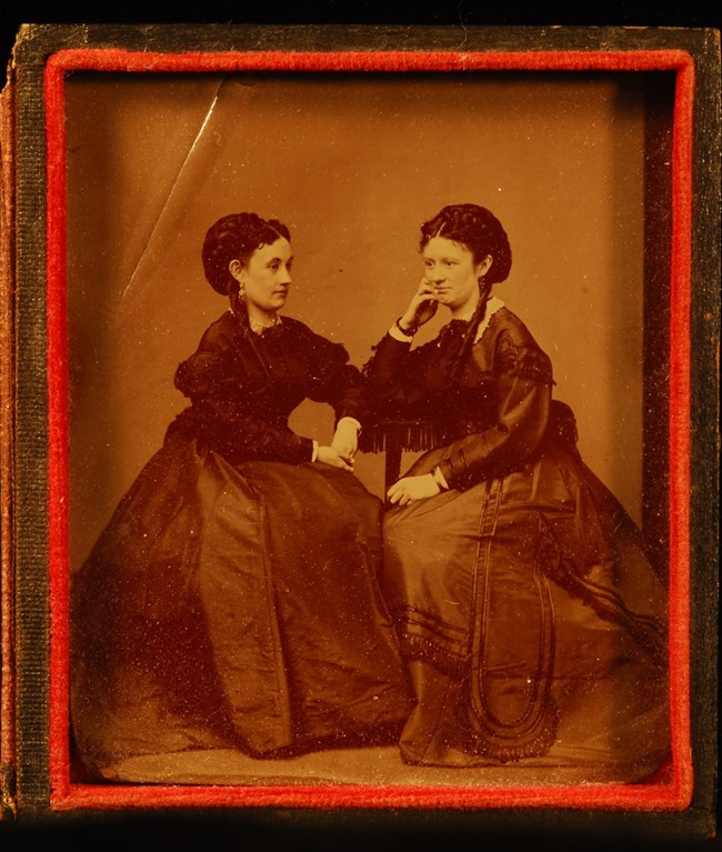 Sepia toned photograph of two women seated in a photography studio in the late 19th century.
