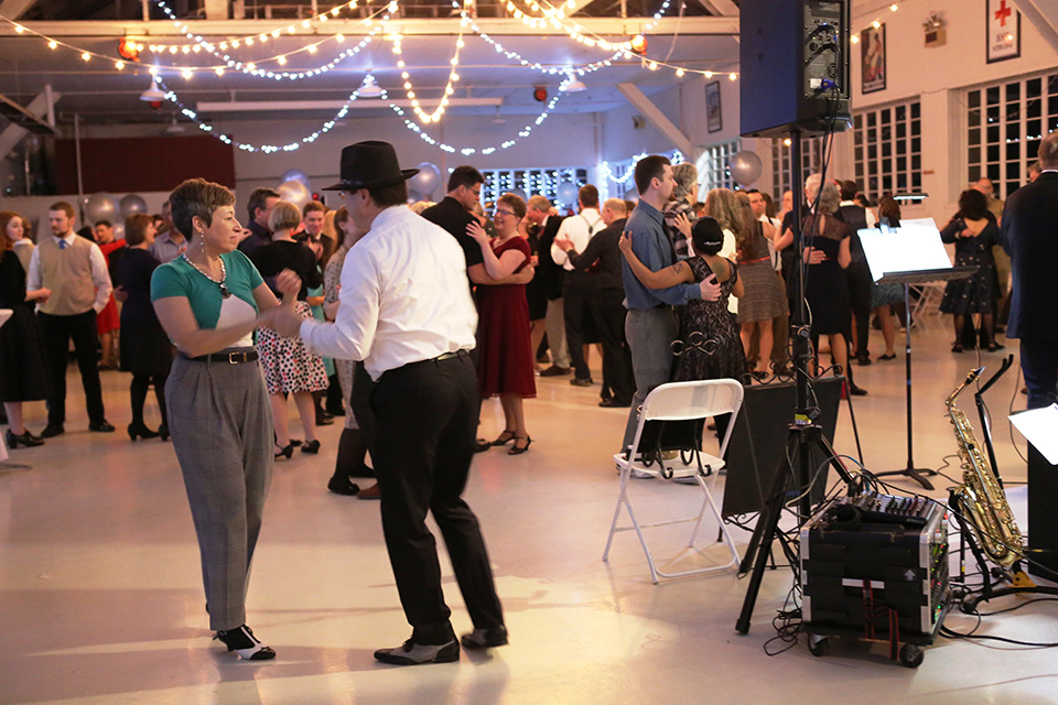 Photo of dancers standing inside dance hall. To the right, part of a big band can be seen. A saxaphone sits on the floor. In the center of the room, many couples dance underneath strings of globe lights.