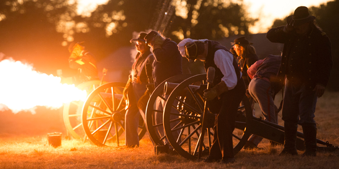 Photo of the firing of a mountain howitzer by volunteers and staff dressed in 19th century Army attire. The photo shows fire belching from the mouth of the cannon, at the exact moment of ignition, while volunteers stand safely beside.